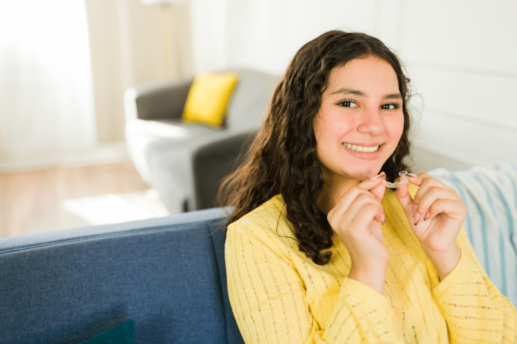 Smiling college-age woman holding a clear orthodontic aligner while seated on a couch