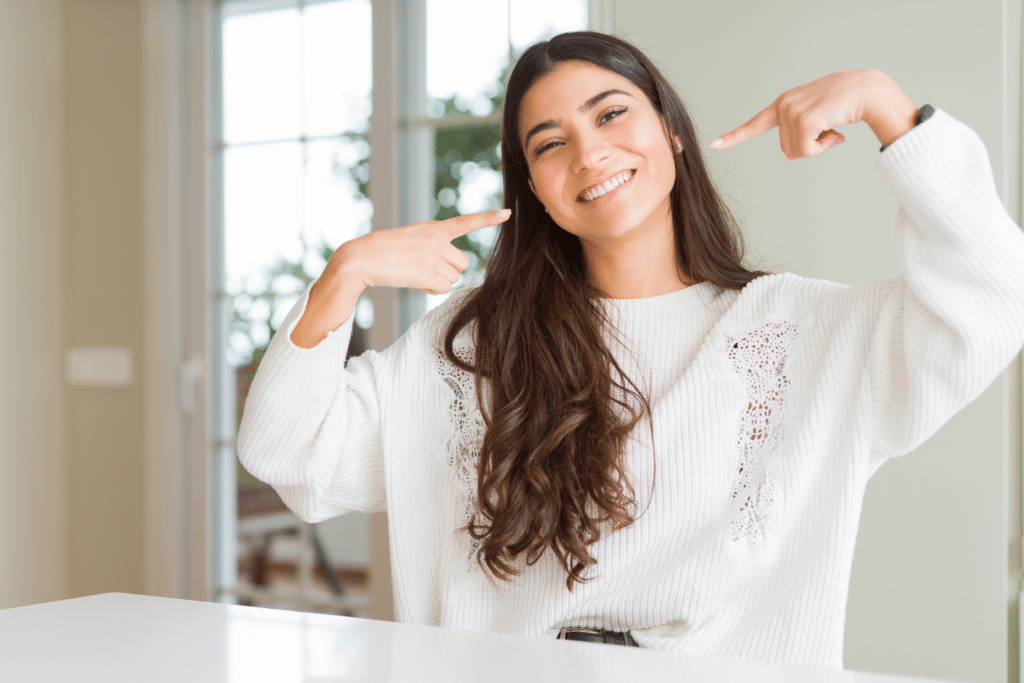 Smiling woman in a white sweater pointing both index fingers at her teeth